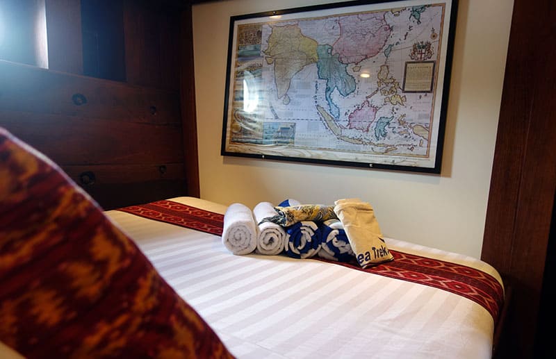 a cabin aboard ombok putih a map of indonesia is hung on a white wall, the bed has a white comforter with red pillows, towels and a bag of gifts sits on the bed