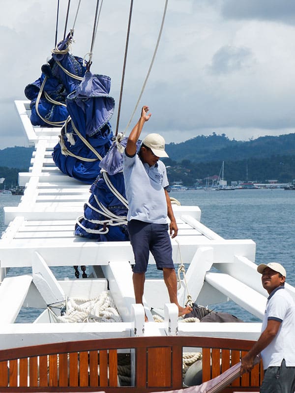 a deck hand oaboard the ombak putih stands on the bow of the white wodden ship holding a rope to the sails