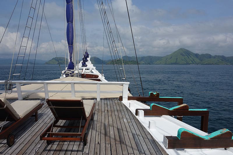 a bow view of the ombak putih, teak lounge chairsface forwards, in the distance a green lush island