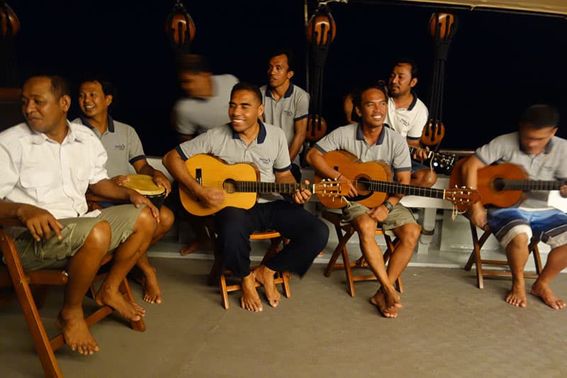 Under the dark sky a group of indonesian crew members sit with guitars and other instruments playing a song aboard a small ship cruise
