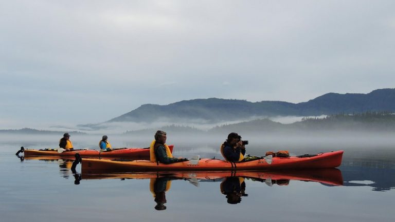 In front of a foggy Alaska mountain range 4 guests wearing life jackets float inside 2 double kayaks and take photos with their camera.