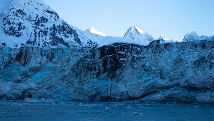 Margerie Glacier with blue ice, black spots & snow-covered mountains in the background on a clear day at sunrise in Glacier Bay National Park.