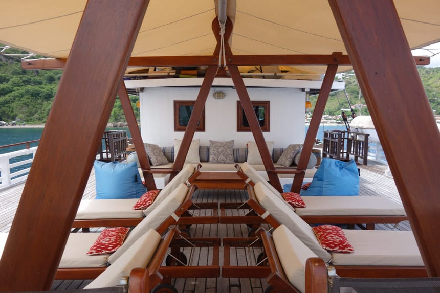 teak beams cross from the ceiling of a overhand to the floor, underneath are lounge chairs with blue and orange pillows