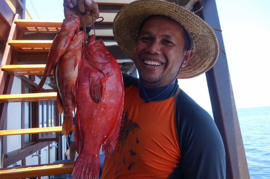an Indonesian guide aboard the ombak putih ship wears a straw hat with a wide brim, he holds a rope with three red fish on the end that he caught for dinner