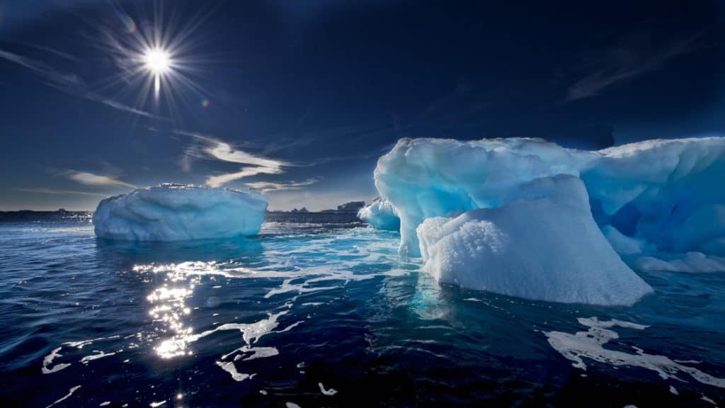 Dark waters & bright blue icebergs float under a bight sun during the Spectacular Ross Sea: West Antarctica Cruise.