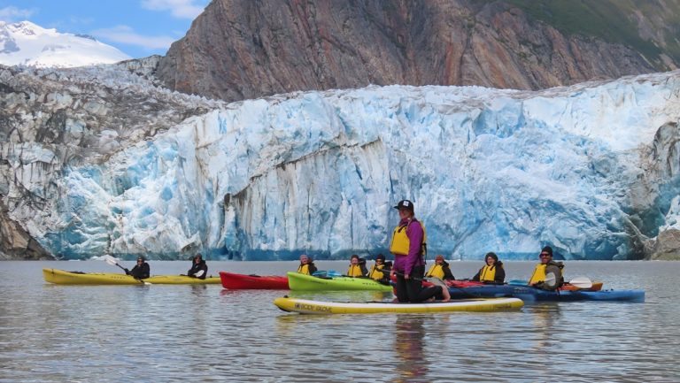 An activity aboard an Alaska cruise, a group of Kayakers and one paddleboarder pose together in front of a large icy glacier.