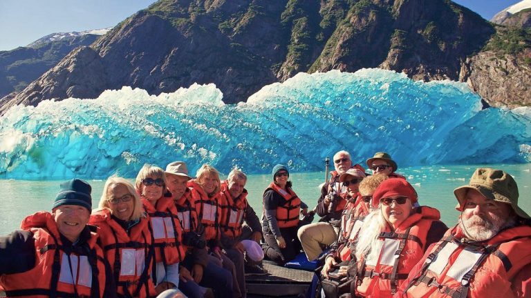 A group of travelers wear orange life jackets as they sit in a zodiac boat in front of a bright teal floating iceberg.