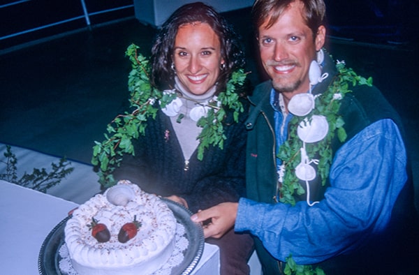 A happy couple smiles and holds their wedding cake off a table, they are celebrating their nuptials on a small ship Alaska cruise. 