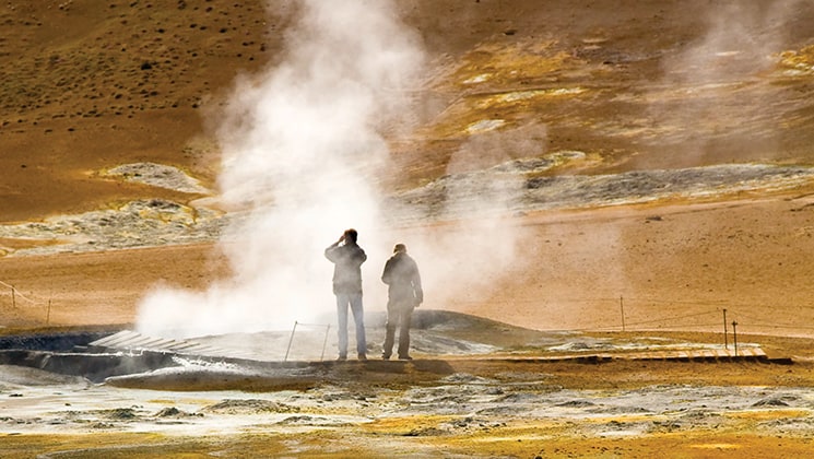 Two travelers stand beside a geyser emitting steam with earthtone rock all around during the Wild Iceland Escape arctic cruise.