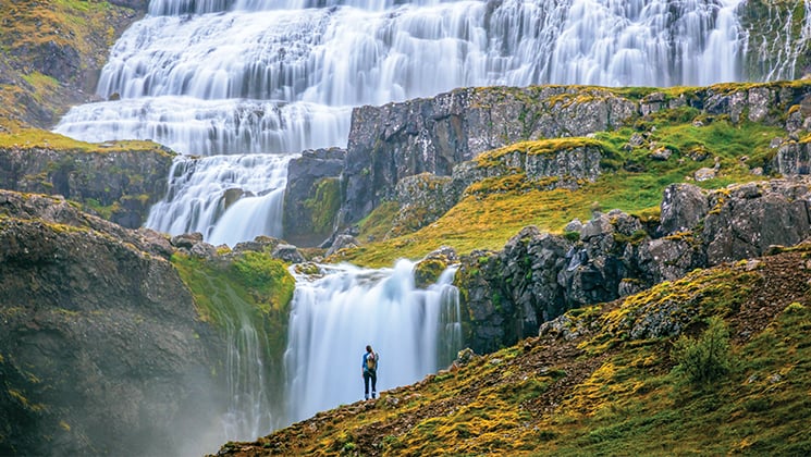 Waterfalls cascade over verdant green rolling hills as a traveler stands in front, during the Wild Iceland Escape arctic cruise.