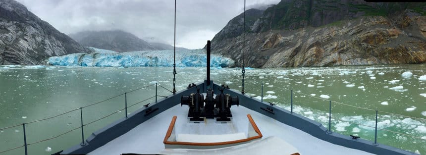 a panoramic photo from the bow of the small Alaskan ship the Westward. Beyond the bow is a teal icy glacier at the end of a fjord seen on an Alaska small ship cruise.