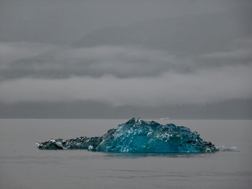 an all grey sky and water are only brought color by a large teal blue iceberg that floats in the water of Alaska's Inside Passage. Seen from the Westward small ship. 
