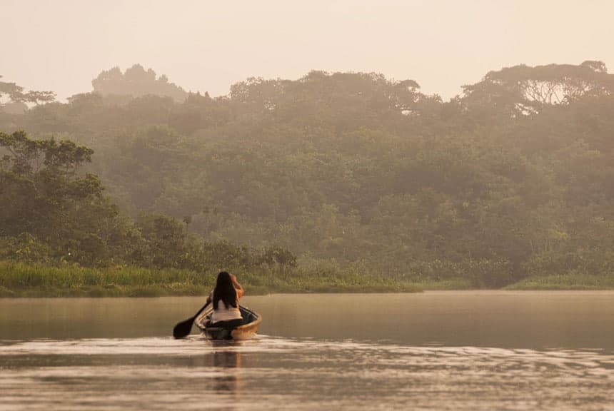  The sun is setting in Ecuador. On Anangu Lake, a woman paddles a wooden canoe away from the camera, it is hazy with a light sepia tone. 