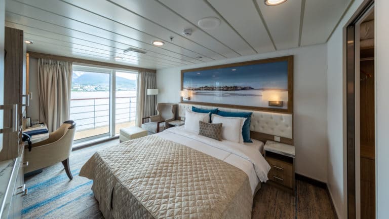 Captain's Suite bedroom aboard Greg Mortimer polar ship, with beige quilted king bed, Antarctica framed photograph & reading lights above headboard, tan chair & sliding glass door to private balcony.