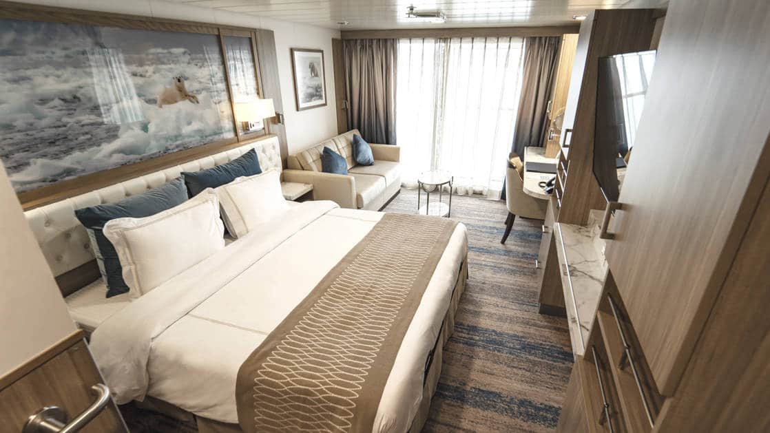 Category A Balcony Stateroom aboard Greg Mortimer polar ship, with white-duvet-covered king bed, marble side table, tan couch, desk, flatscreen TV, wardrobe & sliding glass doors onto private balcony.