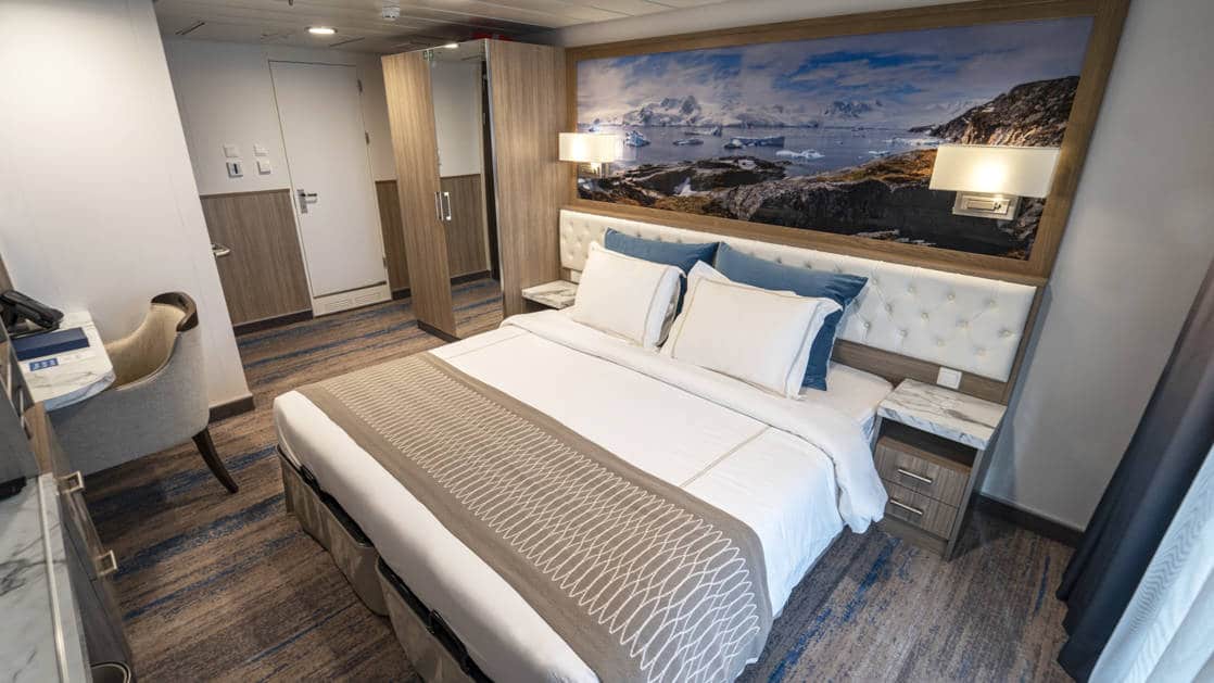 Category B Balcony Stateroom aboard Greg Mortimer polar ship, with white-duvet-covered king bed, marble bedside tables, desk & sliding glass doors onto private balcony.