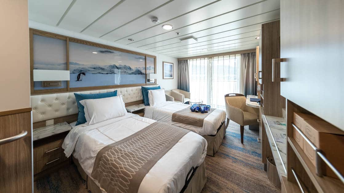 Category C Balcony Stateroom aboard Greg Mortimer polar ship, with 2 white-duvet-covered twin beds, marble bedside tables, tan chair, desk, wardrobe & sliding glass doors onto private balcony.