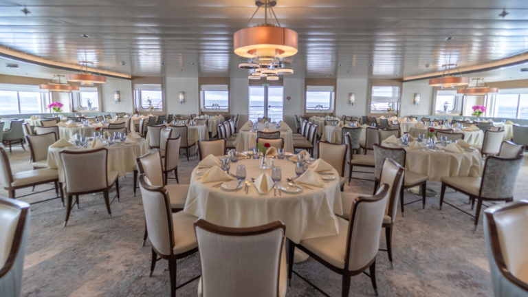 Dining room aboard Greg Mortimer polar ship, with white-tablecloth round tables set for dinner service.