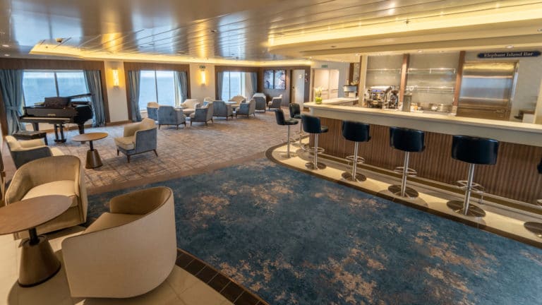 Elephant Island Bar aboard Greg Mortimer polar ship, with beige lounge chairs, grand piano, modern blue barstools & soft lighting.