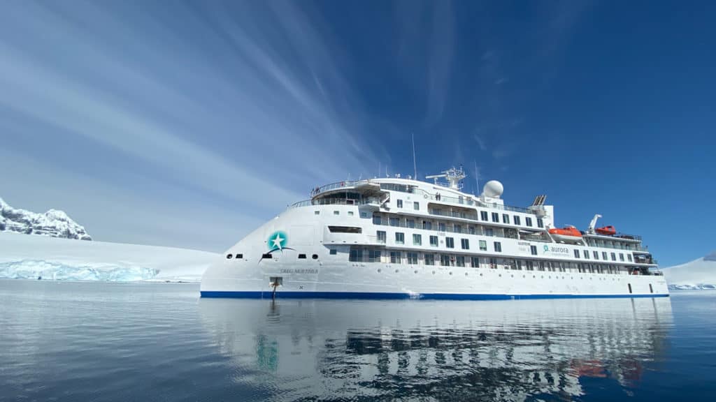 Exterior of Greg Mortimer polar expedition ship, with pointed bow, white exterior & 6 passenger decks, on a sunny day in Antarctica.