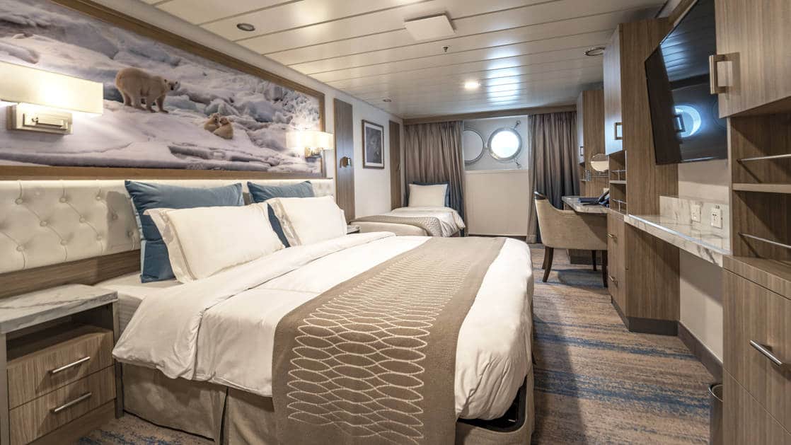Triple Stateroom aboard Greg Mortimer polar ship, with white-duvet-covered king bed & twin bed, marble bedside tables, tan chair, desk, flatscreen TV, wardrobe & porthole.