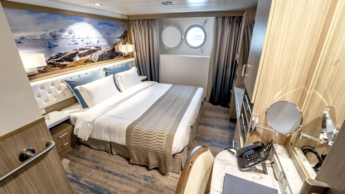 Twin Stateroom aboard Greg Mortimer polar ship, with white-duvet-covered king bed, marble bedside tables, tan chair, desk, wardrobe & porthole.