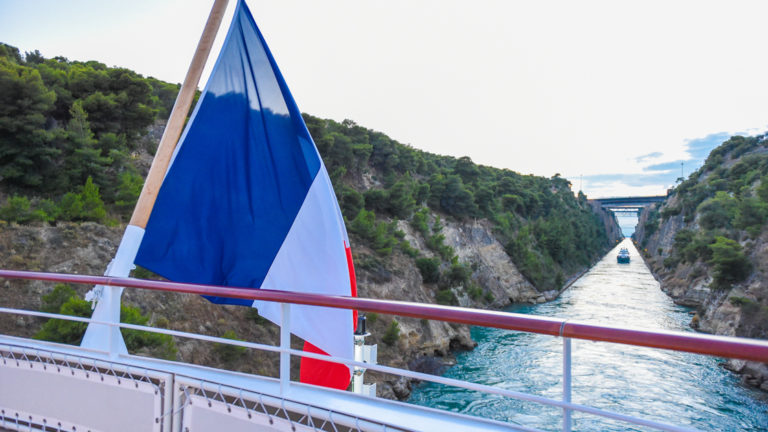A French flag flies from the aft upper deck of a luxury small ship as it cruises through the narrow Corinth Canal, lined by greenery and tall rock walls, during the Best of Croatia Cruise.