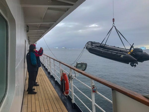 from deck four of hebridean sky polar expedition ship, the ships railing surrounds a path, outside of it a black zodiac raft is being lifted and let down into the water