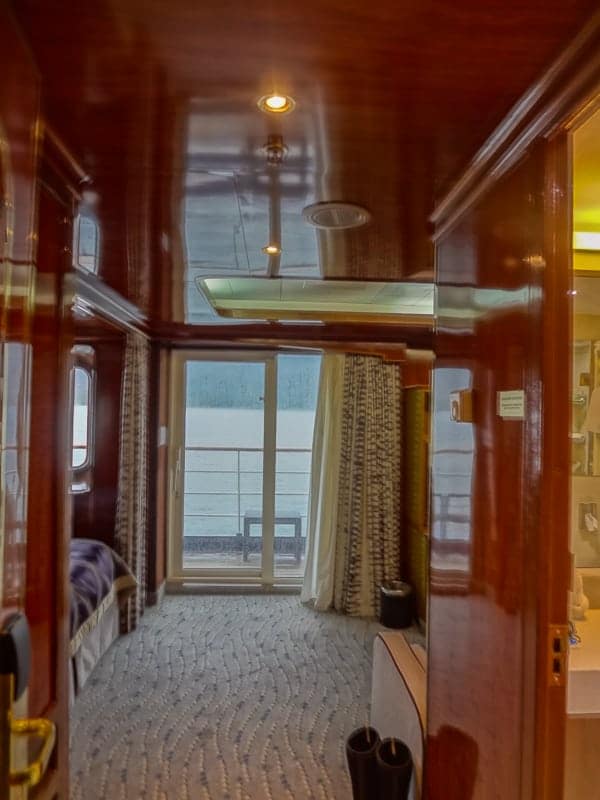 a rich wooden suite cabin aboard hebridean sky polar expedition ship shows the end of the bed and a sliding glass door out to a private balcony. 