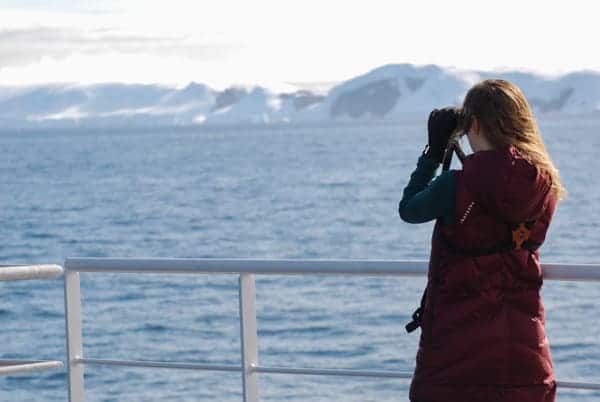 from the deck of hebridean sky polar expedition ship , a female guest faces out toward the sea with binoculars to her eyes looking for birds