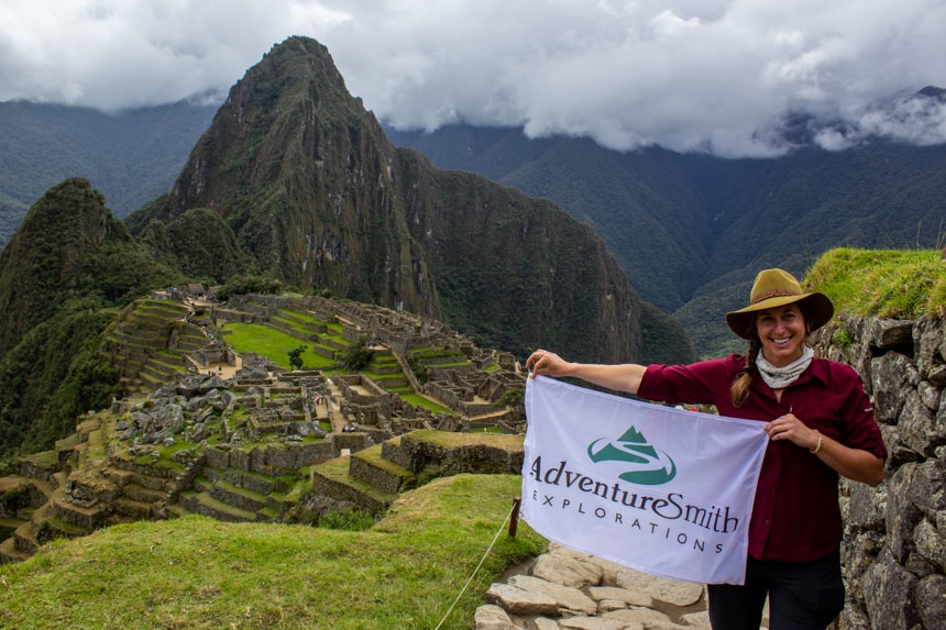 A female traveler stands in front of Machu Picchu, towering green cliff sides with ancient walled town below her as she holds a white AdventureSmith Explorations flag. 