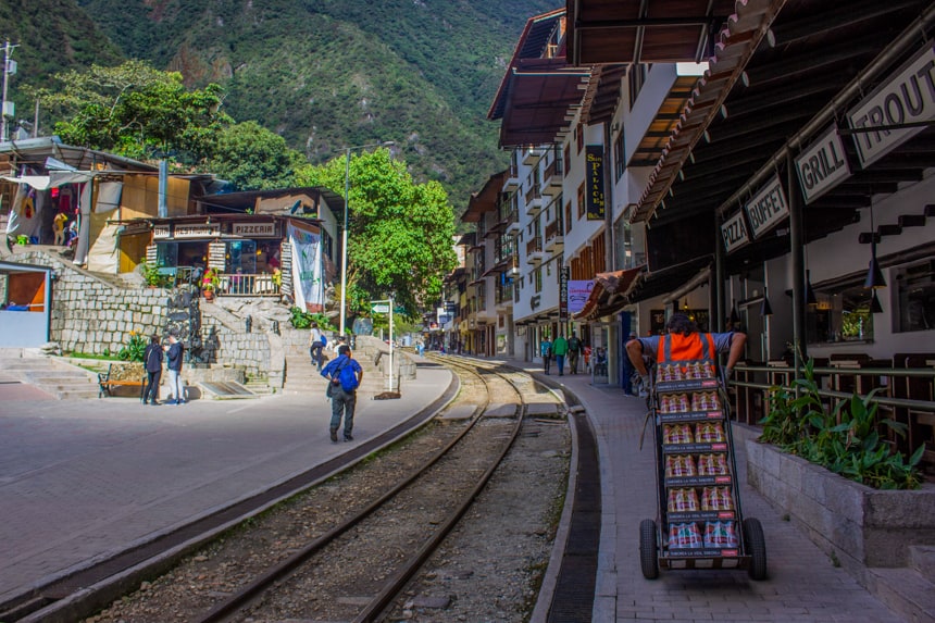 Downtown in Aguas Calientes, Peru. A view from the train tracks that split the city buildings. People are walking around the streets infront of large green mountainside