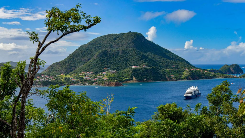 Small expedition ship with dark blue hull & white upper decks sits in Caribbean waters near lush, green, mountainous islands.
