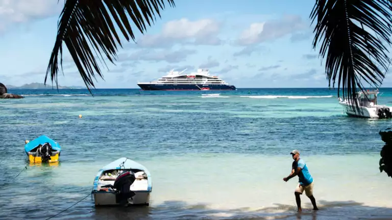 A man walks toward a dinghy on a white-sand beach with palm tree fronds above & a luxury ship in the distance during the Pearls of the Caribbean luxury cruise.