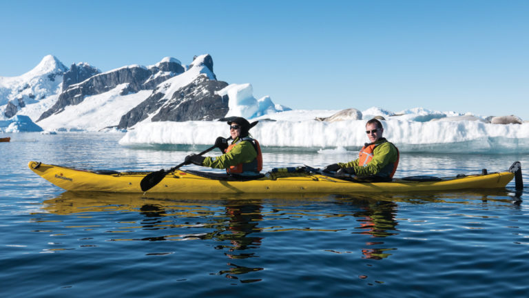 Tandem kayakers in a yellow boat paddle in calm waters with snow-covered mountains in the background, on a sunny day during the Spirit of Antarctica expedition.