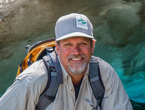 AdventureSmith founder Todd Smith stands infront of a teal glacier wearing an orange backpak and AdventureSmith logo hat