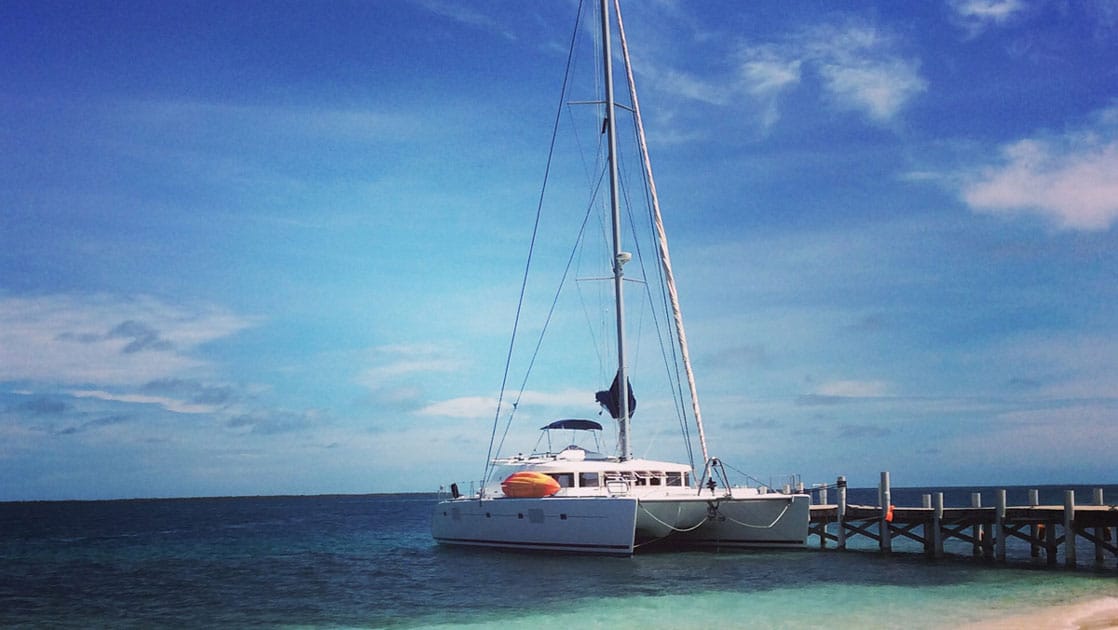 White catamaran Sand Star sits docked in turquoise water by a white-sand beach on a sunny day in Belize.