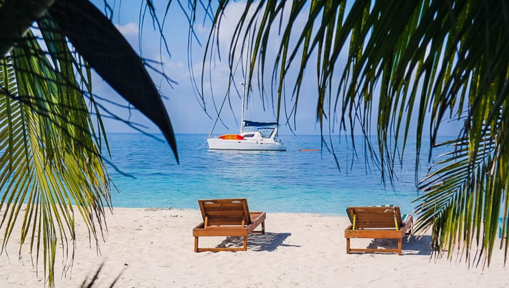 Two empty wooden chaise loungers sit on a white-sand beach with palms swaying & a Belize catamaran charter cruise boat sitting offshore.