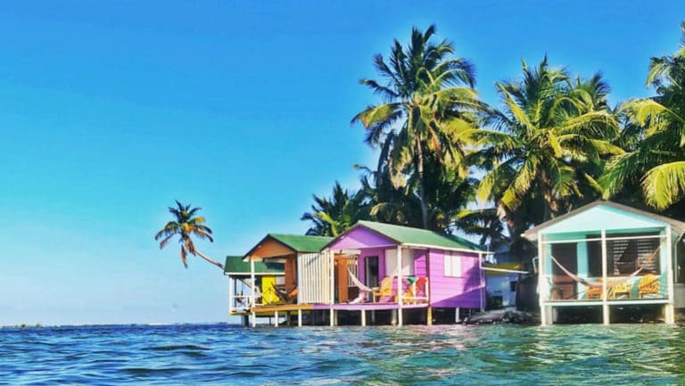 Colorful beach houses sit on stilts above clear blue waters with palm trees above, on a sunny day.