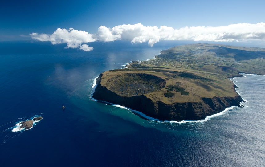 an aerial shot of Easter Island, a green land mass island that sits in the green ocean and matching blue sky