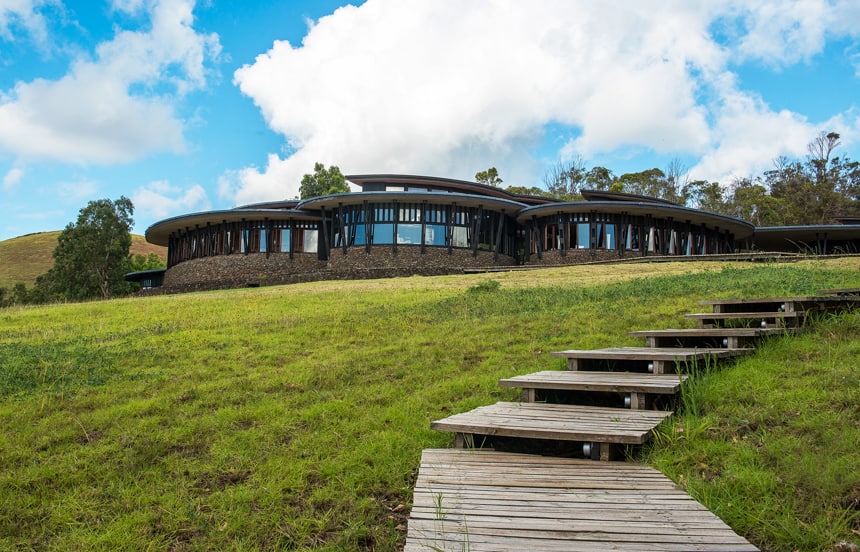 Explora Rapa Nui Lodge, a circular stone buildings with wrap around windows sit on a grassy hisiude witjh wooden steps reaching to it.