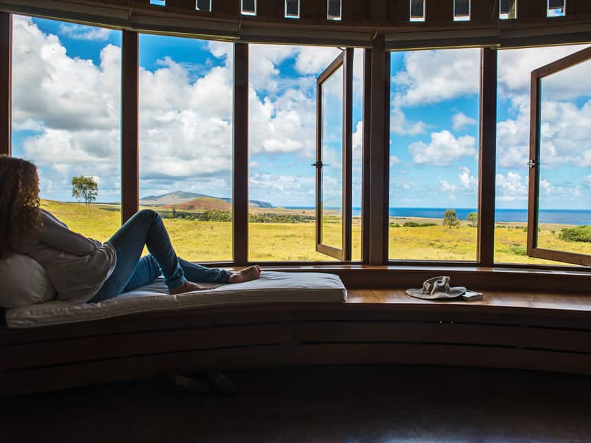 at Explora Rapa Nui Lodge, a female travelers lays on a bench seat infront of windows that open to the outside, a view of a green meadow and blue ocean in the distance