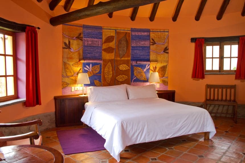 A room inside Sol Y Luna a Peru Lodge inside the Sacred Valley, orange walls with red window covers, a white queen bed sits agains peruvan wall art