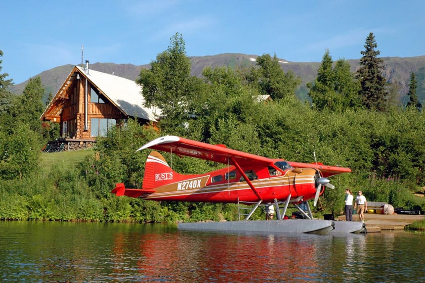 A bright red float plane sits in front of the dock at Winterlake Lodge in Alaksa behind it is lush forest with the lodge sitting ontop of the hilllside