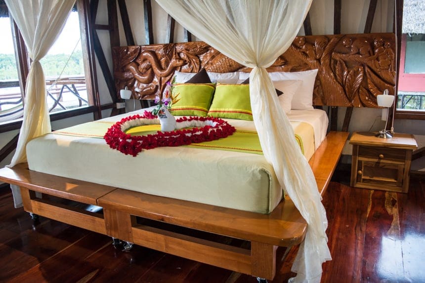inside a cabana at the Napo Wildlife Center, a king bed with a heart made of flowers sits infront of a wodden cardev headboard surrounded by a bug net