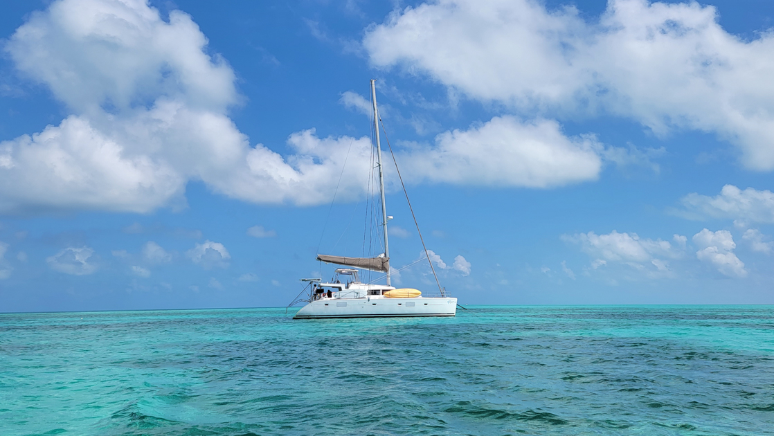 Discover Belize catamaran with white exterior & stowed sails sits in turquoise water above dark reef on a sunny day.