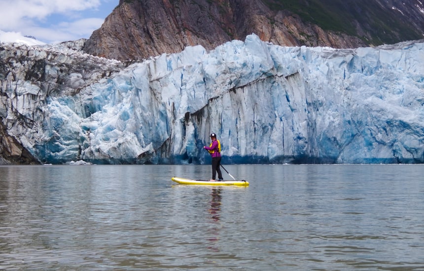 a female traveler wearing a yellow life jacket and pink long sleeve sweater, paddles on a stand up paddle board infront of a Glacier on a small ship cruise in Alaska.