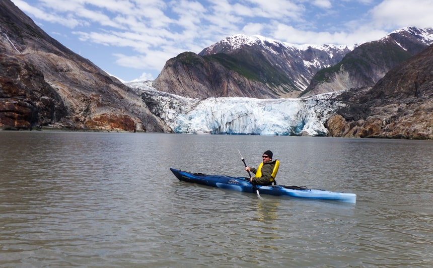 A single kayaker wearing a yellow life jacket paddles a blue kayak in front of a glacier on a blue sky day in Alaska on the islands whales and glaciers cruise.
