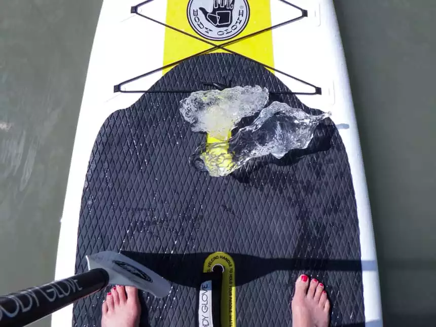 Shot from above, you see someones feet standing ontop of a white and black paddle board that is floating in water, two crystal pieces of ice are ontop with the end of a paddle.