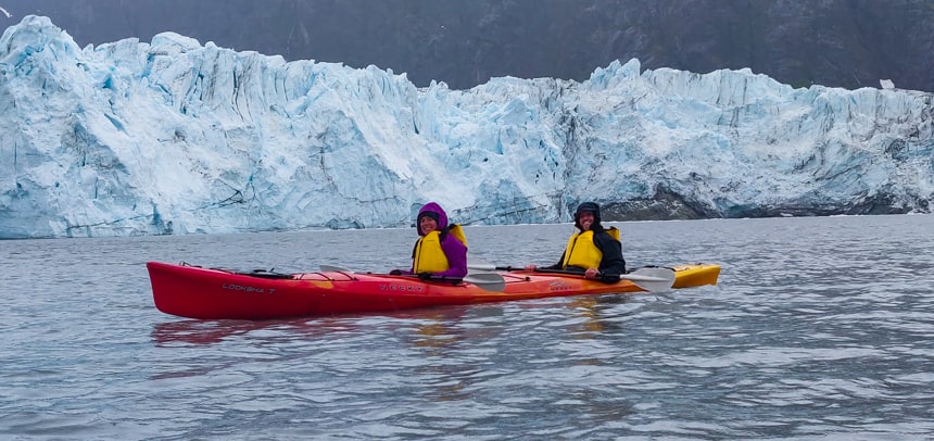 two happy guests paddle a red tandem kayak behind them is a jagged teal and white glacier, an activity done on Islands whales and glacier small ship cruise